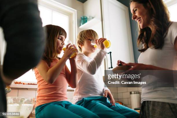 mother looking at children drinking juice while sitting on kitchen counter - orange juice stockfoto's en -beelden