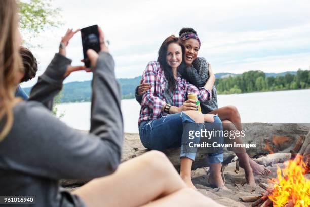 woman photographing happy female friends sitting on tree trunk by campfire against river - dosen schießen stock-fotos und bilder