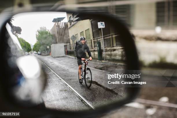 male commuter riding bicycle on wet street seen through side-view mirror of car - specchietto di veicolo foto e immagini stock