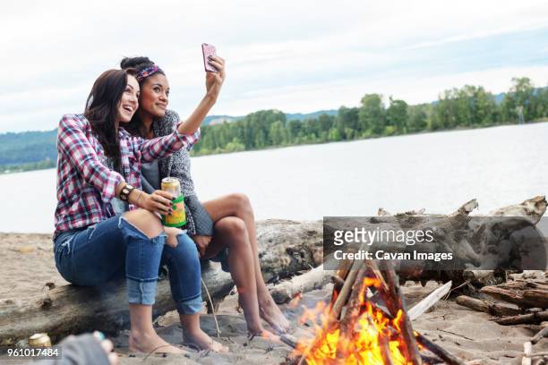 female friends taking selfie while sitting on tree trunk by campfire against river - dosen schießen stock-fotos und bilder