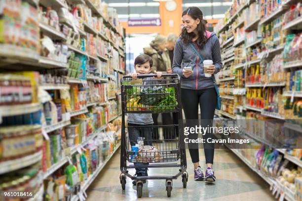 woman walking by son carrying shopping cart at supermarket - paar gruppierung stock-fotos und bilder