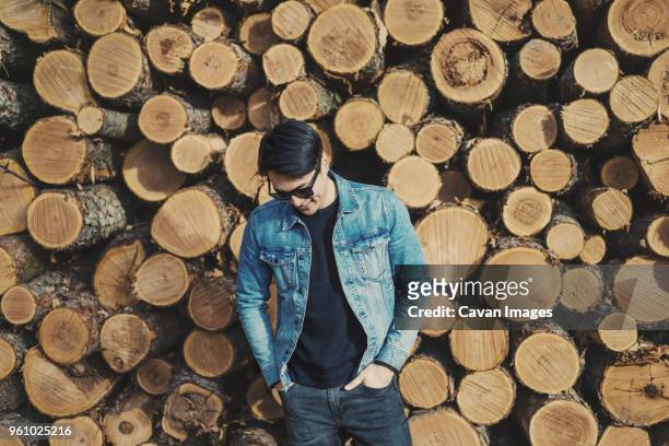 young man with hands in pockets standing against stack of logs - デニムジャケット ストックフォトと画像