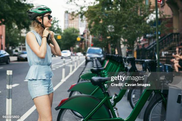 smiling woman wearing helmet while standing by bicycles on street - bike sharing stock-fotos und bilder