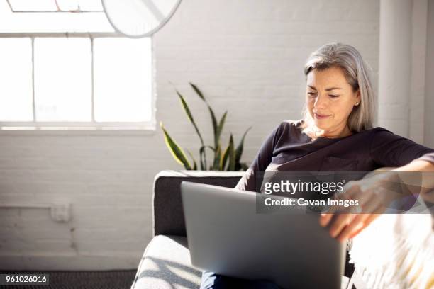 woman using laptop while sitting on sofa in living room at home - notebook stockfoto's en -beelden