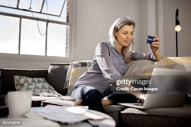 mature woman holding debit card while using laptop on sofa - baby boomer home stock pictures, royalty-free photos & images