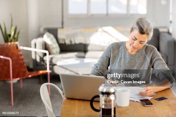 concentrated woman writing on paper while working on laptop in living room - 50s woman writing at table stock pictures, royalty-free photos & images