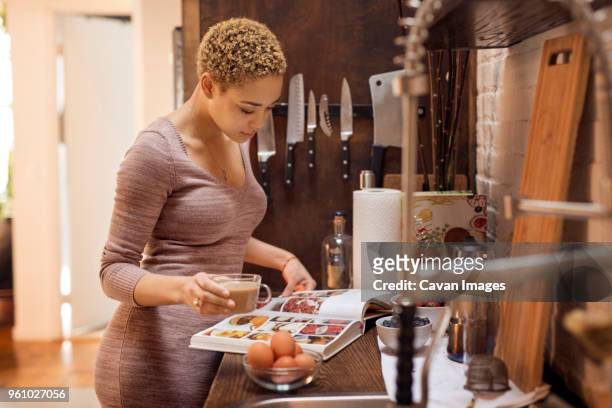 woman reading recipe book while holding tea cup in kitchen - reading cookbook stock pictures, royalty-free photos & images