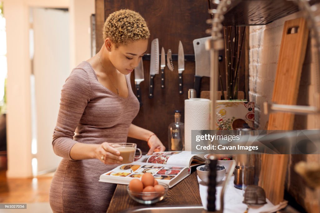 Woman reading recipe book while holding tea cup in kitchen