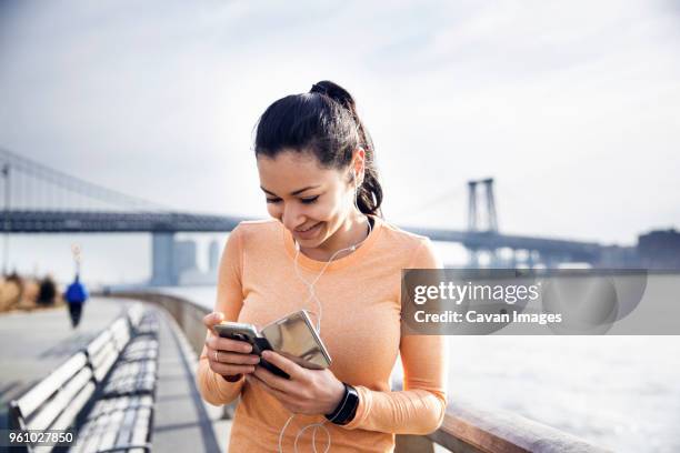 happy female athlete listening music through smart phone with williamsburg bridge in background - suspension training stockfoto's en -beelden