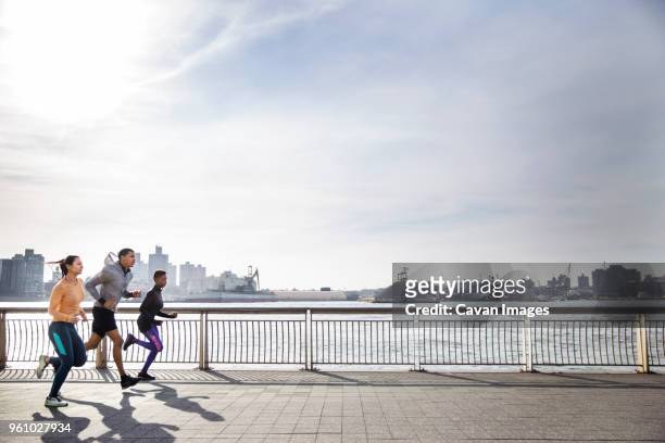 side view of determined athletes running on footpath by river against sky - jogging city stock pictures, royalty-free photos & images