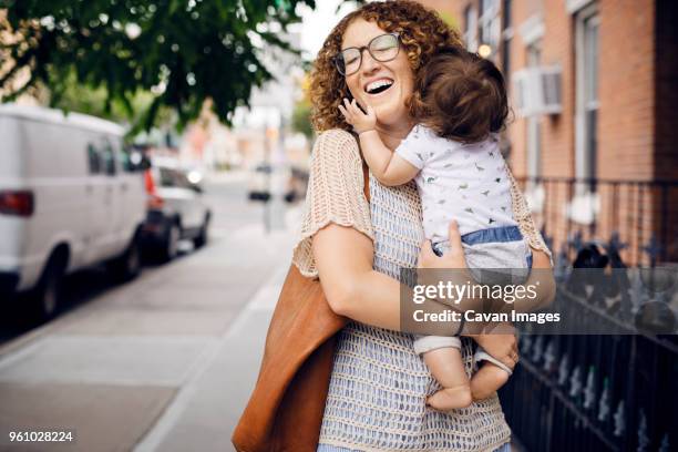 cheerful mother carrying baby boy on footpath in city - brown purse stock-fotos und bilder