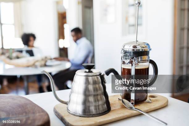 french press and kettle on cutting board with couple in background - coffee plunger stock pictures, royalty-free photos & images