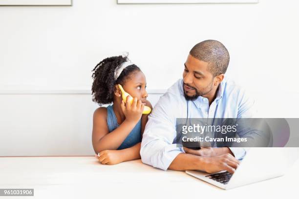 man holding mobile phone looking at daughter while sitting at table - banana phone stock pictures, royalty-free photos & images