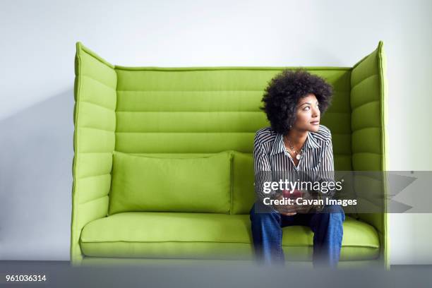 businesswoman looking away while sitting on sofa against wall in office - capelli neri foto e immagini stock
