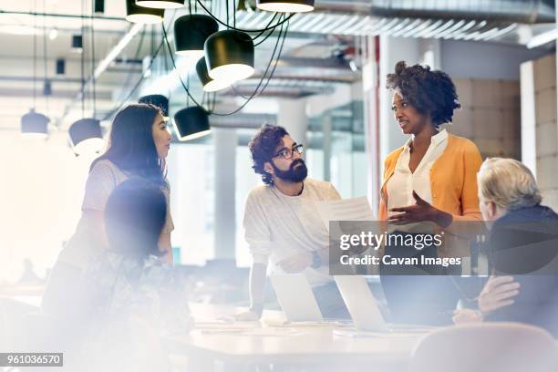 business people looking at female colleague explaining documents while standing at conference table - business partner light stock-fotos und bilder