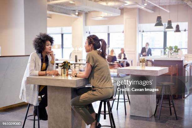 businesswomen talking while sitting in cafeteria at creative office with colleagues in background - pause café bureau photos et images de collection