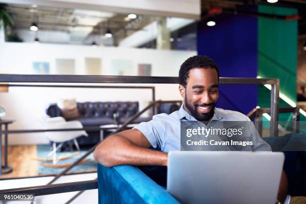 smiling young businessman using laptop while sitting on sofa at office - young man laptop couch photos et images de collection