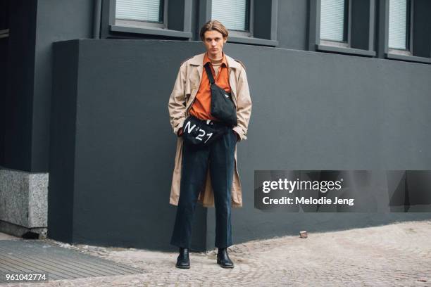 Model Jai Piccone wears a tan trenchcoat, orange shirt, Prada cross-body bag, a N21 Numero Ventuno bag by his waist, black pants, and black shoes...