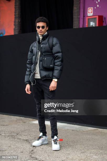 Model Joel Leon Perreira uses earphones in one ear and wears round sunglasses, a black Polo puffer jacket, black Nike cross-body bag, gold watch,...