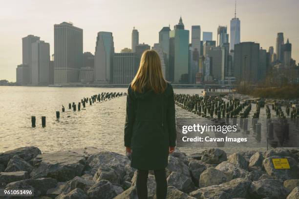 rear view of woman standing on rocks by river in city against sky during sunset - blonde hair from behind stock pictures, royalty-free photos & images