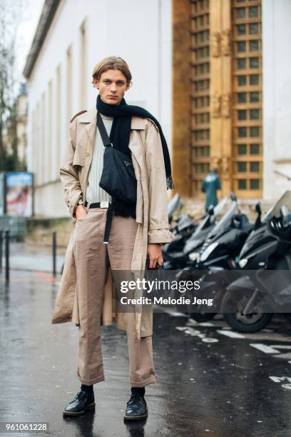 Model Jai Piccone wears a tan trenchcoat, tan pants, black shoes, a black Prada bag across his chest, black scarf, and holds black headphones during...