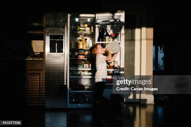 side view of boy drinking milk from bottle while standing against open refrigerator in darkroom at home - drinking milk stock pictures, royalty-free photos & images
