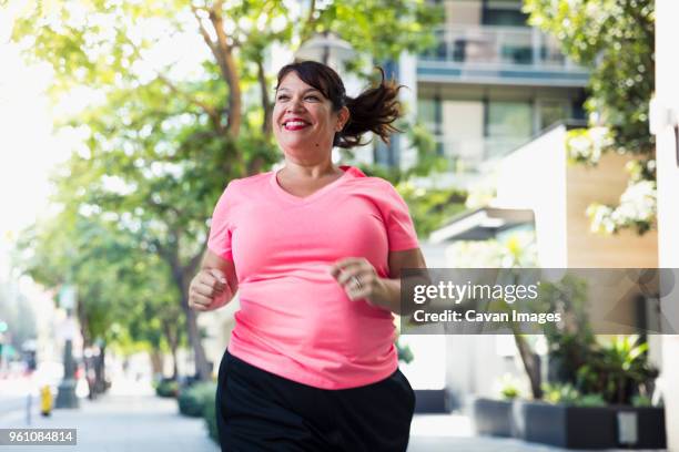 cheerful woman jogging on footpath in city - overweight stockfoto's en -beelden