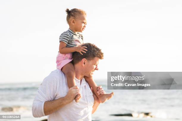 father carrying daughter on shoulders while standing at beach against clear sky - genderblend2015 stock pictures, royalty-free photos & images