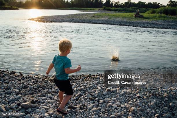 rear view of boy throwing stones in lake during sunset - boy throwing stock-fotos und bilder