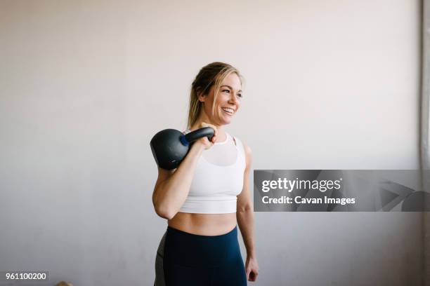 smiling female athlete carrying kettlebell while standing by wall in gym - kettlebell stockfoto's en -beelden