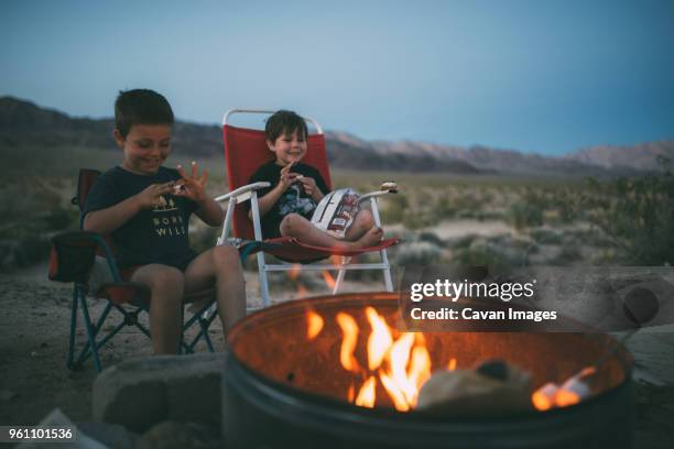 happy brothers eating marshmallows while sitting on chairs at campsite during dusk - joshua tree national park california stock pictures, royalty-free photos & images