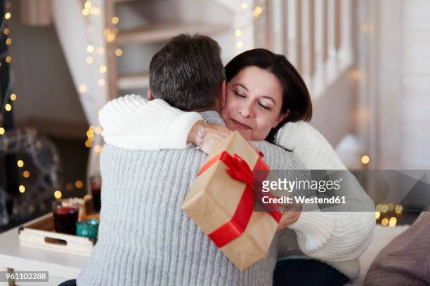 happy mature couple with gift box hugging in living room - holiday gift stock-fotos und bilder