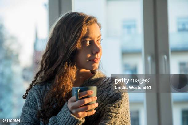 portrait of teenage girl with coffee mug looking out of window in the evening - ringellocke stock-fotos und bilder