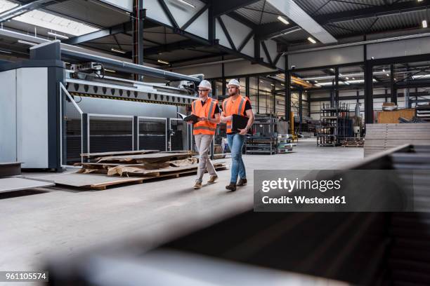 two men wearing hard hats and safety vests walking on factory shop floor - produktionshalle stock-fotos und bilder