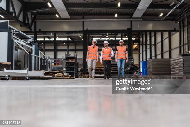 three men wearing hard hats and safety vests walking on factory shop floor - naderen stockfoto's en -beelden