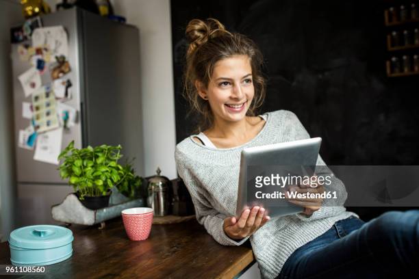 smiling woman sitting at table holding tablet - woman kitchen stock-fotos und bilder