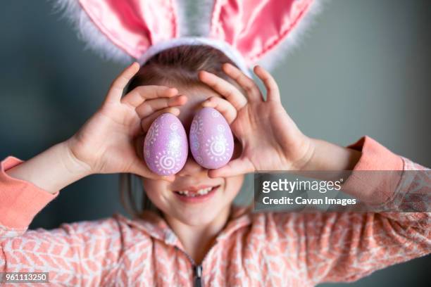 playful girl wearing rabbit ears headband while holding easter eggs at home - rabbit costume stock pictures, royalty-free photos & images