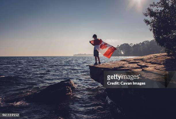 full length of boy holding canadian flag while standing on cliff against sea and sky during sunny day - canada flag stock pictures, royalty-free photos & images