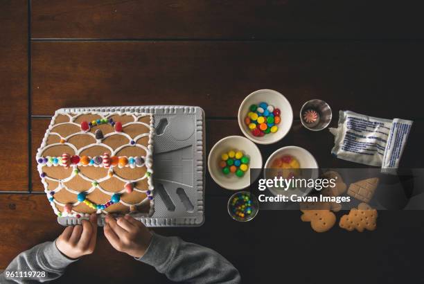 cropped hands of boy making gingerbread house on wooden table during christmas at home - gingerbread house stock pictures, royalty-free photos & images