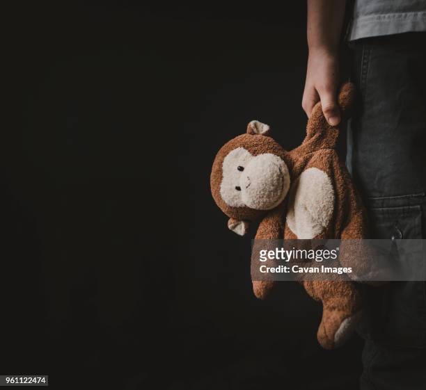 cropped image of boy holding stuffed toy while standing against black background - stuffed toy stock-fotos und bilder
