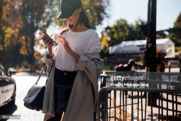 Blanca Padilla checks her phone and wears a baseball cap/"dad hat," jacket off her shoulders, white top, black jeans, and a black Chanel purse after...