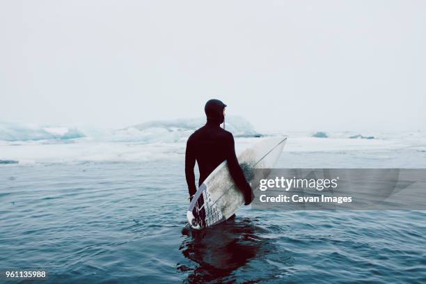 man carrying surfboard while standing in icy sea against clear sky during winter - man hauling stock pictures, royalty-free photos & images