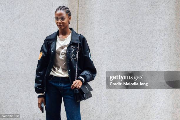 Model Karly Loyce wears thin-frame circular glasses, a black Fear of God varsity jacket, Bob Marley t-shirt, YSL black bag, and blue jeans during New...