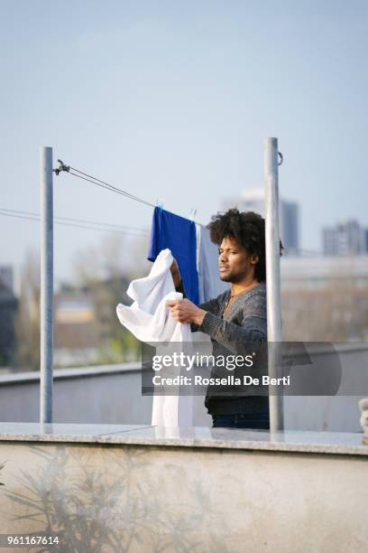 young beautiful man hanging up laundry on rooftop terrace - afro man washing stock pictures, royalty-free photos & images