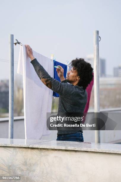 young beautiful man hanging up laundry on rooftop terrace - afro man washing stock pictures, royalty-free photos & images