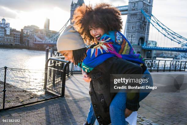 young giving young woman piggyback outdoors, tower bridge in background, london, england, uk - london fashion fotografías e imágenes de stock