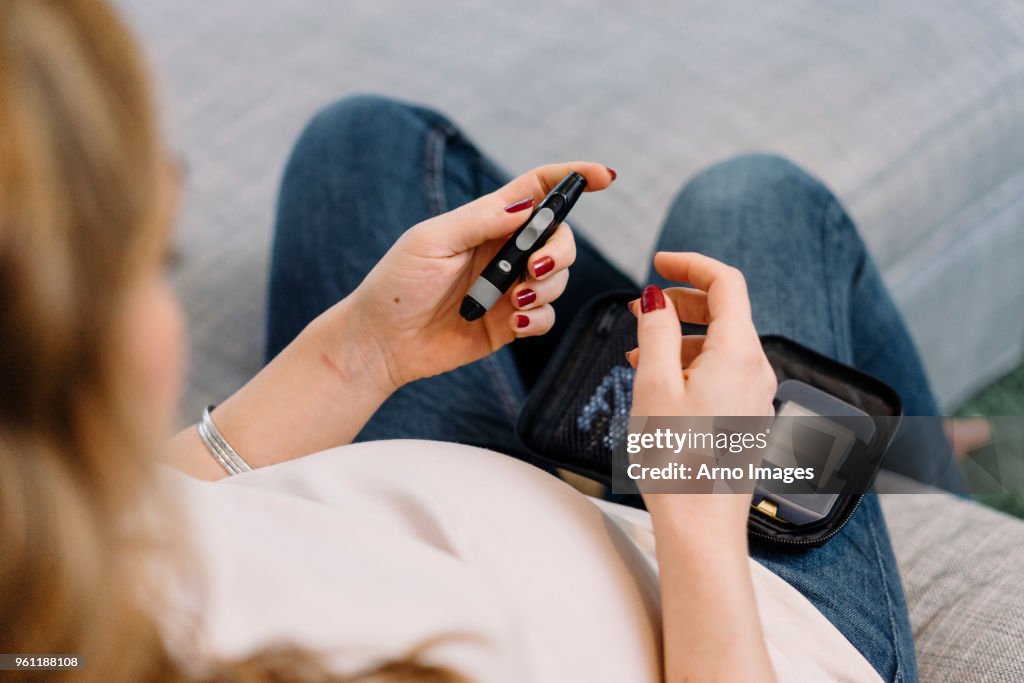 Pregnant woman on sofa taking blood sugar test, over shoulder view