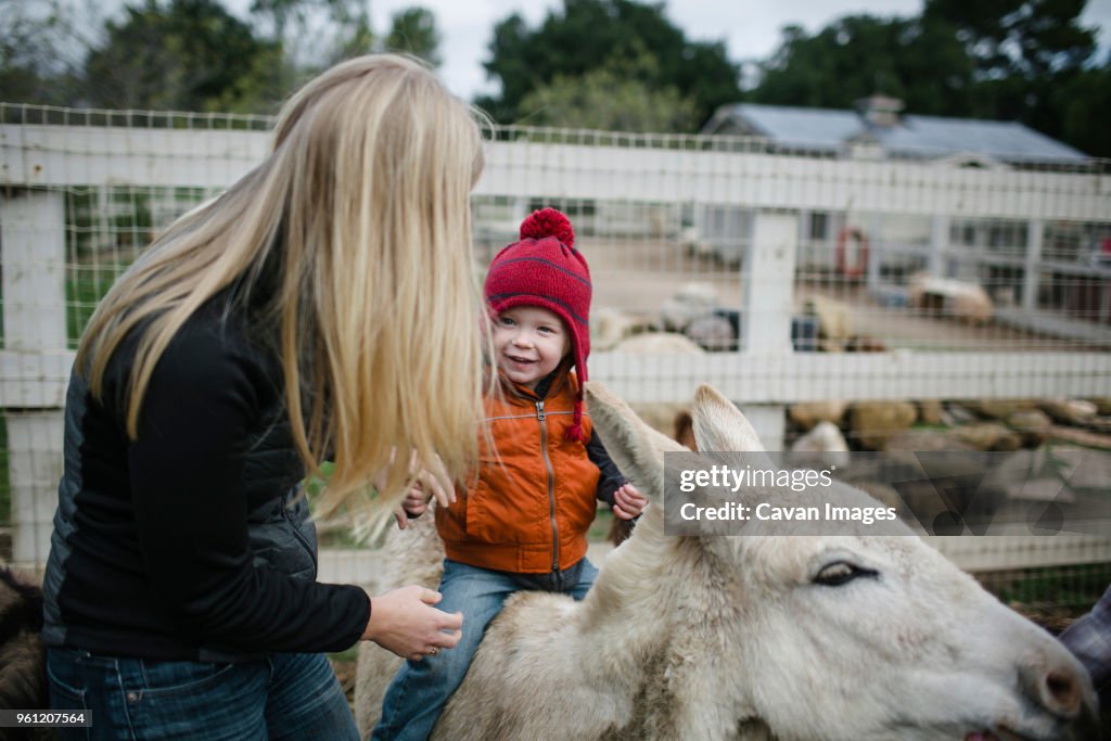 Side view of mother standing by son sitting on donkey at farm