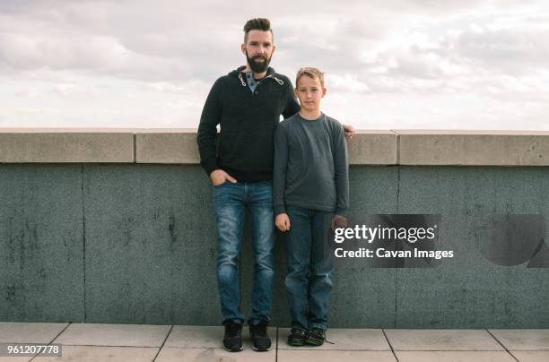 portrait of father with son standing by retaining wall against cloudy sky - genderblend2015 stock pictures, royalty-free photos & images