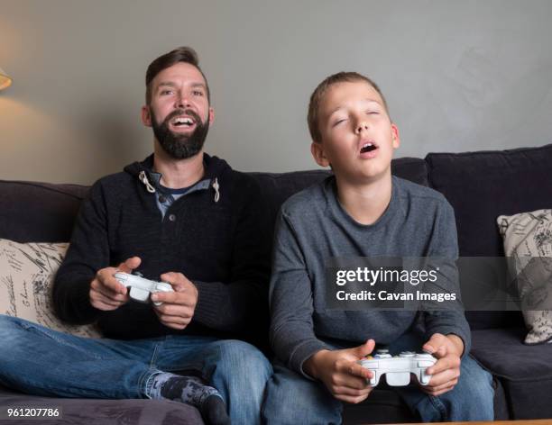 father with son playing video game while sitting on sofa at home - genderblend2015 stock pictures, royalty-free photos & images